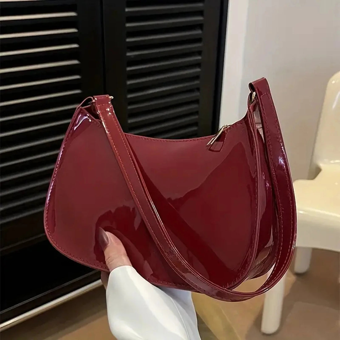 Bolso baguette burdeos de polipiel de charol brillante sujeto por el asa delante de unas contraventanas oscuras