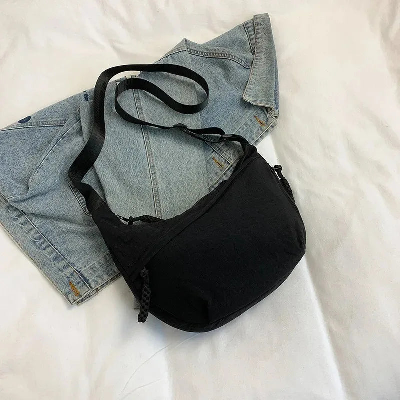 Bolso bandolera de nailon negro apoyado sobre una cazadora vaquera en una cama blanca