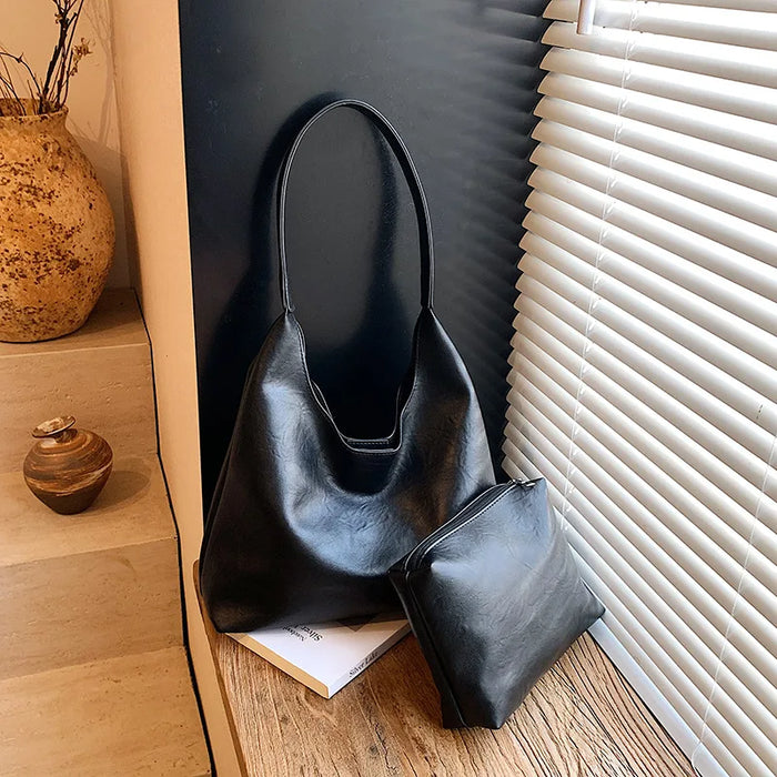 Bolso hobo negro de polipiel de pie sobre un suelo de madera junto a una persiana, con su neceser a juego al lado