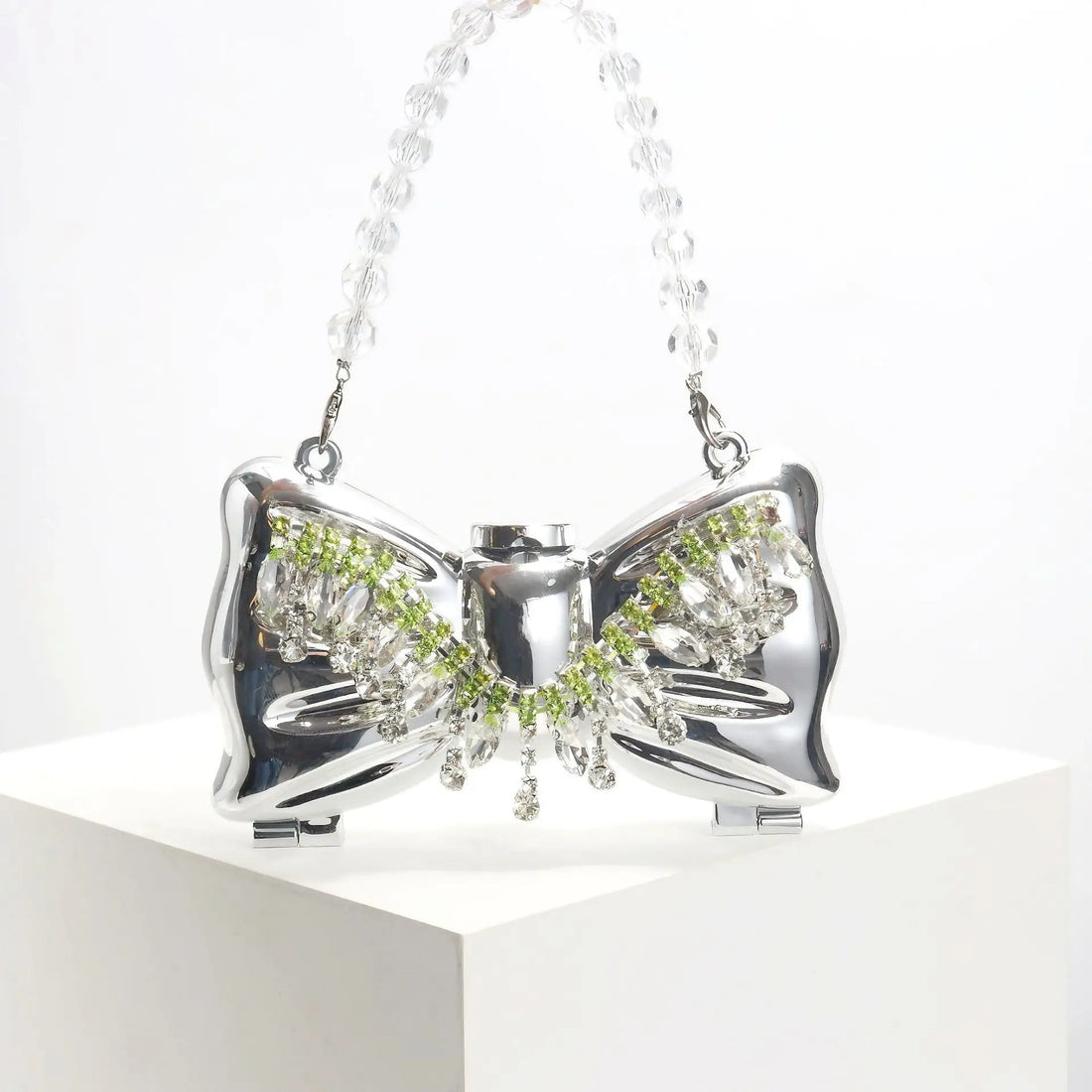 Mini clutch plateado con forma de lazo sobre un pedestal blanco, con el asa transparente efecto cristal